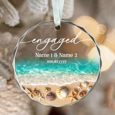 Custom Beach Engagement Glass Ornament – Engagement Christmas Gift with a beach and ocean wave design, featuring the word “Engaged,” personalized couple names, and a special date, tied with a cream ribbon and displayed beside daisies and a gift box.