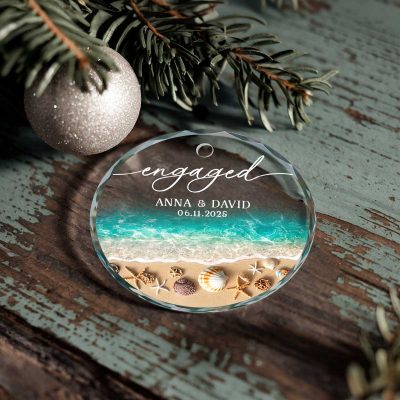 Custom Beach Engagement Glass Ornament – Engagement Christmas Gift with a beach and ocean wave design, featuring the word “Engaged,” personalized couple names, and a special date, tied with a cream ribbon and displayed beside daisies and a gift box.